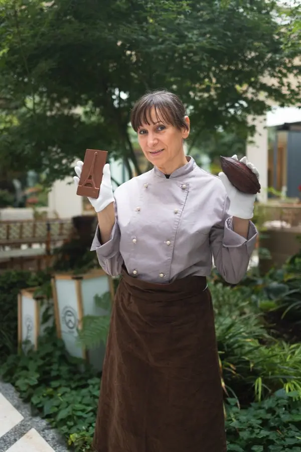 Ana Moreira en tenue de chocolatière dans un patio d'hôtel aux allures champêtres, tenant d'une main une cabosse de cacao, et de l'autre main une tablette Tower artisanale.